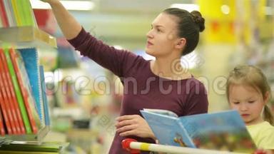 在超市里微笑的母亲和女儿。 女儿帮助母亲选择一本书。 可爱的女儿坐在里面